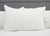 A large, white Downlite Primaloft Down Alternative Pillow with a smooth surface rests on a white bed, against a light gray upholstered headboard. A fabric care tag adds to the clean, minimalistic look.
