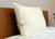 The Down Etc. Aquaplush Pillow, a large and fluffy white pillow with a smooth cover, stands upright against a wooden headboard on a neatly made bed with a white comforter. A green plant is visible to the right.
