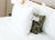A close-up of a bed with crisp white sheets and a large Sysco Guest Supply Dream Escape Down Alternative Pillow (formerly Dream Essence) in front. Behind it, a green floral decorative pillow with a care tag peeks out from under the white pillow.