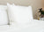 A clean, minimal bedroom features a white bed with Pacific Coast Feather Tria Down & Feather Pillows by Pacific Coast Feather Company—used at many Ritz-Carlton® Hotels—plus fluffy bedding and a green potted plant on a light nightstand.