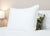 A large white Sysco Guest Supply Dream Escape Down Alternative Pillow sits upright on a bed with white sheets. Behind it is a smaller pillow with a green, black, and gold floral pattern. A lit bedside lamp is partially visible on the left.