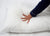 A hand with red-painted nails presses on the center of a plush Downlite Primaloft Down Alternative Pillow, which compresses to show its hypoallergenic comfort. The person wears a dark blue long-sleeve shirt with white trim.