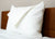 A Pillowtex Bamboo Pillow Cover in white, zipped over a large pillow, rests upright against a wooden headboard. A folded white sheet lies on top, and the bed features a clean, padded mattress for an inviting look.