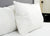 A Pillowtex White Goose Down & Feather Pillow (75% feather/25% down) from Pillowtex rests upright against a matching pillow on a neatly made bed with white sheets, a gray headboard, and dark furniture near the door.