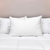A Pillow Factory Downlite 75/25 Waterfowl Feather/White Down Pillow is centered on a neatly made bed with a white comforter and two white pillows, set against a clean, minimal backdrop with a soft, light-colored wall.