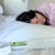 A dark-haired woman in a red-and-white shirt rests on a bed, using Keeco’s Choice Hotels® Soft and Firm Polyester Pillow Combo Pack; a green "Choice Hotels" tag is visible, with the background gently blurred.